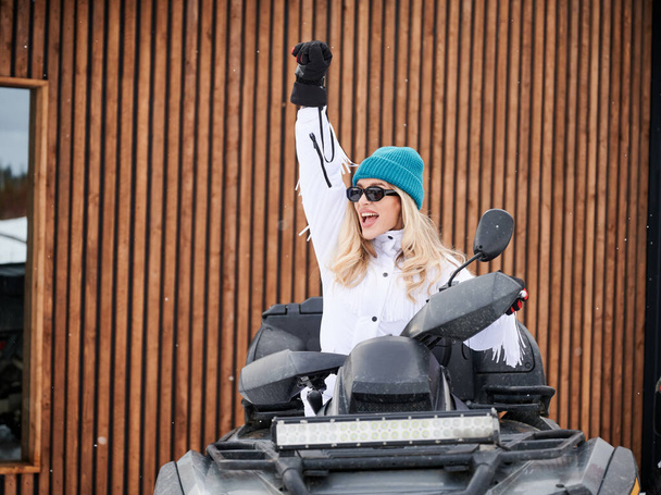 Portrait of beautiful woman posing on offroad four-wheeler ATV with wooden house on background. Concept of active leisure and winter activities. - Photo, Image