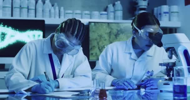 Scientist, chemists or medical engineers using a microscope to test samples and analyze data while writing down their results. Two colleagues researching a virus and its cure in a uv lit lab. - Πλάνα, βίντεο