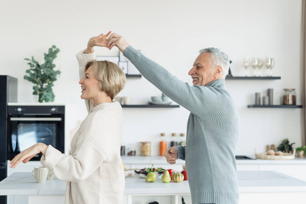 Married family couple smiling and dancing slowly in kitchen with modern interior and cozy atmosphere. Elderly man and woman falling in love, dating, enjoying life and spending free time together - Foto, Imagen