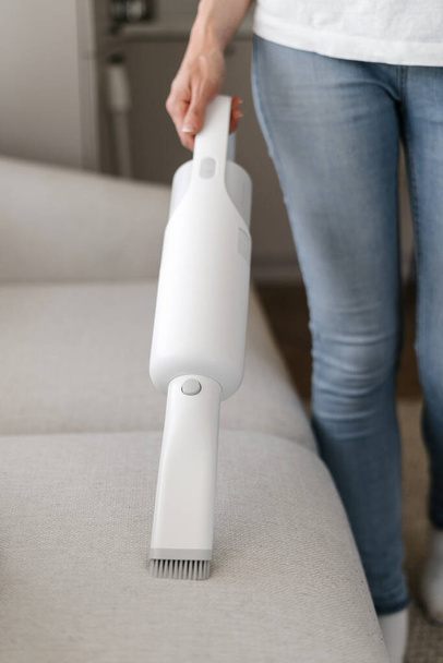 Cropped view of woman using wireless vacuum to clean sofa in living room. Concept of cleanliness. Household equipment usage. Housewife chores. Cleaning service worker - Photo, image