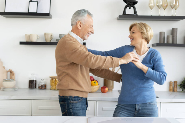 Happy and mature couple celebrate the anniversary of marriage, dancing together at kitchen. Senior man looking at his wife with love, spending family holidays at home, having fun, enjoying moment - Foto, immagini