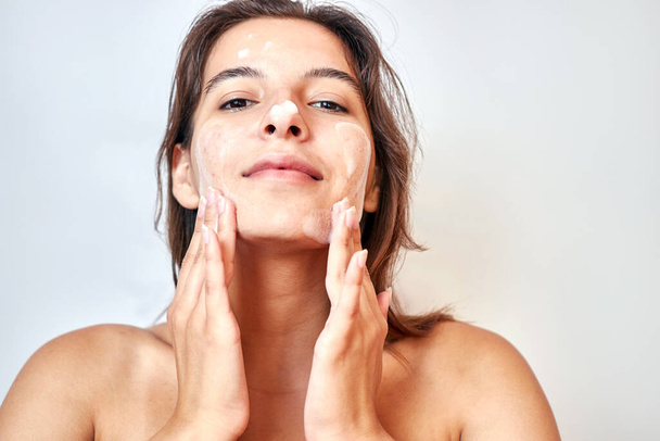 Uma menina aplica espuma no rosto para limpar a pele. Conceito de cuidado facial. - Foto, Imagem