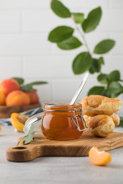 A jar of apricot jam with a spoon inside, ripe apricots in a wooden plate and a branch with green leaves on a light background. Preparations for the winter, fruit preservation. - Foto, afbeelding