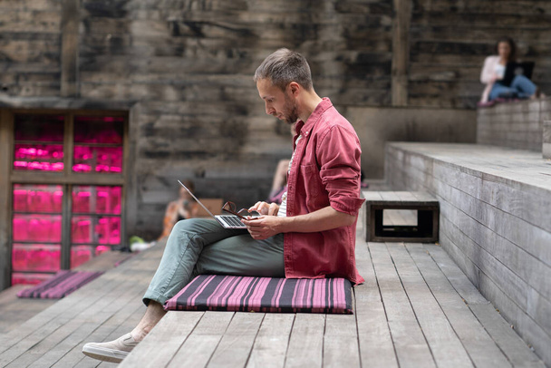 Focused concentrated man using laptop outside, doing online freelance job. Pensive European student guy studying outdoors, sitting on wooden bench. Working spaces for freelancers and self-employed - Foto, Bild