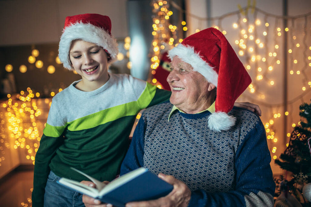Isoisä ja pojanpoika istuvat tuolilla ja lukevat kirjaa joulun aikana.  - Valokuva, kuva