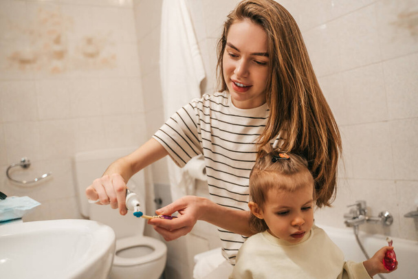 Smiling young woman squeezing the toothpaste from the tube on the toothbrush of her child - Photo, image