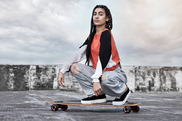 Unbeatable on the board. Cropped portrait of an attractive young female skater crouching on her longboard on a rooftop against a stormy backdrop - 写真・画像