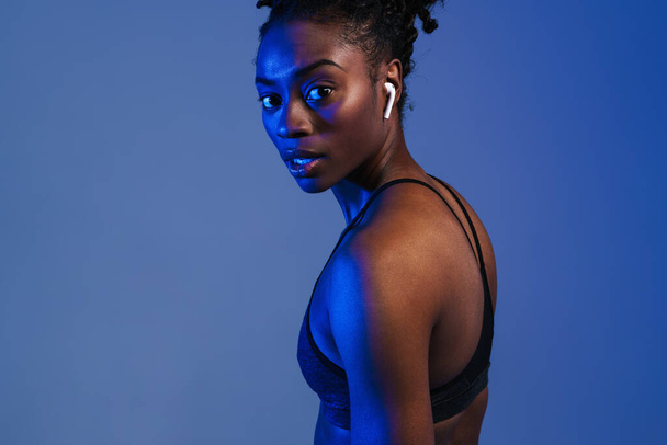 Black young sportswoman in earphones looking at camera isolated over blue background - Фото, зображення