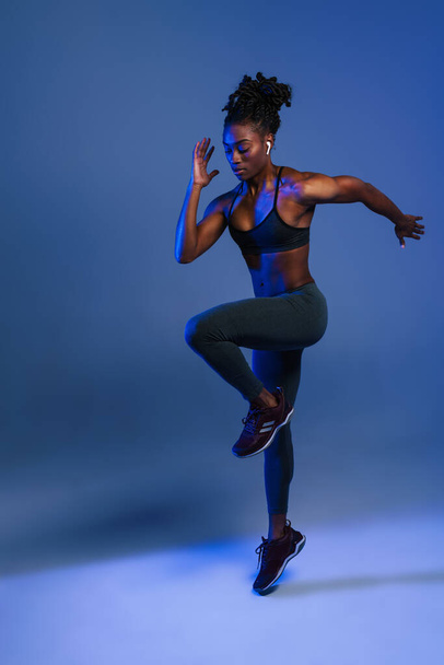 Black sportswoman in earphones doing exercise while working out isolated over blue background - 写真・画像