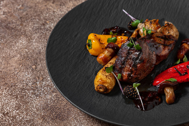Deer filet mignon steak. Grilled steak with vegetables, paprika, chilli, mushrooms. The food is topped with a sauce made from young cone jam. The food lies on a black, round, slate plate. The plate stands on a dark, fabric background, coffee color. - Photo, image