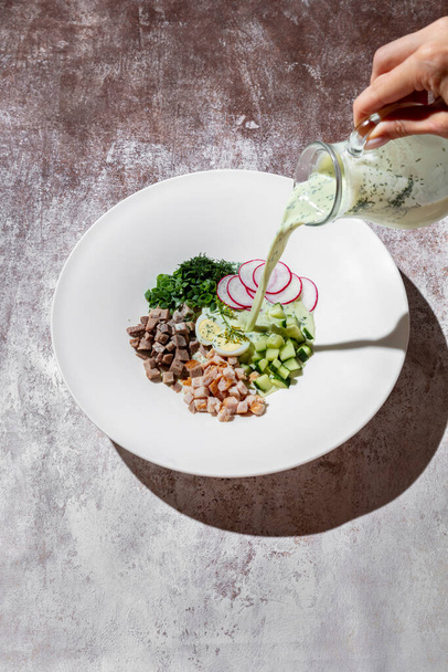 Pieces of pork, chicken with egg, cucumber, onion, radish and parsley in a plate on a gray background with a ruy that sprinkles the whole plate with okroshka in a jug - Photo, Image