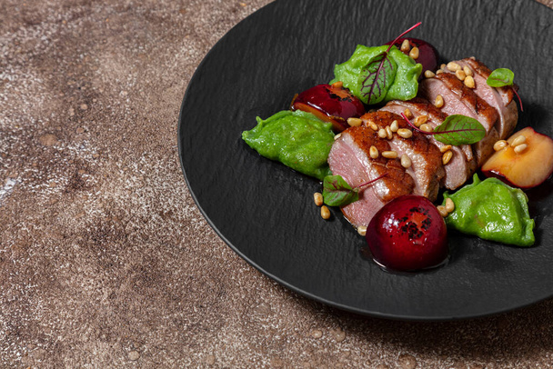 Duck breast in plum sauce, grilled and cut into portions. Topped with pine nuts and lettuce leaves. Nearby are basil ravioli with mashed potatoes and porcini mushrooms. Plum sauce and caramelized plums lie side by side. The food lies on a black, slat - Fotoğraf, Görsel