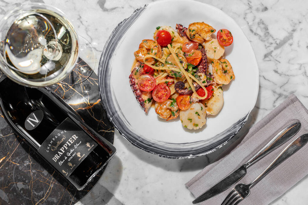 Pasta with seafood. Homemade pasta with scallop, octopus, shrimp and cherry tomatoes in a creamy herb sauce. Pasta lies in a round, light ceramic plate. The plate stands on a light marble table. Nearby is a glass of prosecco, cutlery on a linen napki - Photo, Image