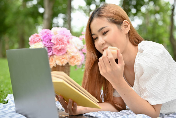Atrakcyjna młoda Azjatka leżąca na kocu piknikowym, czytająca książkę, jedząca słodycze i mająca miły piknik w ogrodzie. - Zdjęcie, obraz