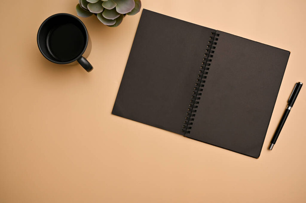 Minimal orange beige workspace background with empty black spiral notebook, pen, a cup of coffee and decor plant. top view - Zdjęcie, obraz