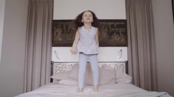 Small caucasian brunette girl jumps on the bed. Happy childhood concept. Play in the morning at home. Wake up in the morning and dance at home. High quality 4k footage - Filmagem, Vídeo