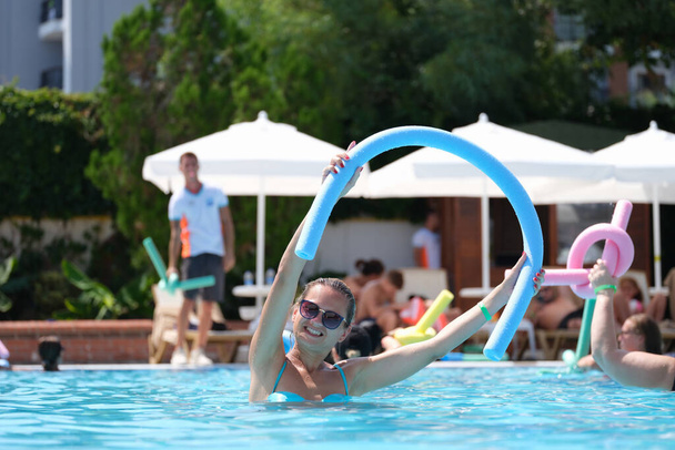 Atrakcyjna uśmiechnięta kobieta w okularach kąpielowych z makaronem w basenie. Aerobik wodny i fitness w basenie na wakacjach w hotelu - Zdjęcie, obraz