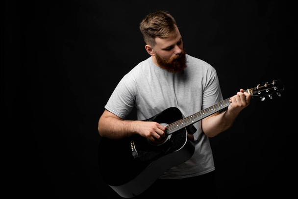 Concentrated brutal handsome bearded man in grey t-shirt playing acoustic guitar. Portrait of a male singer and guitarist - Foto, Imagem