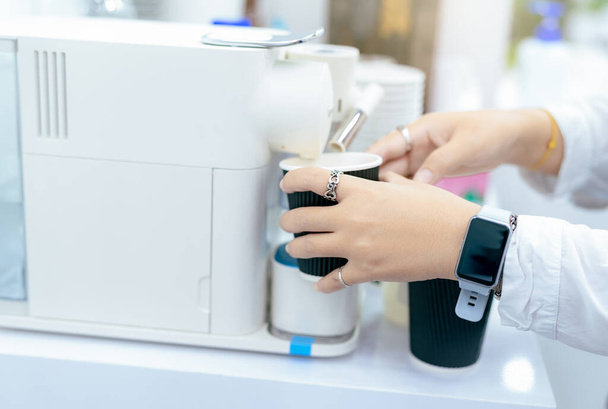 Woman making a cup of hot coffee with capsule coffee machine. Woman holding black cup taking coffee from a capsule coffee machine on the table. Espresso maker. Morning drink. Modern home equipment. - Foto, afbeelding