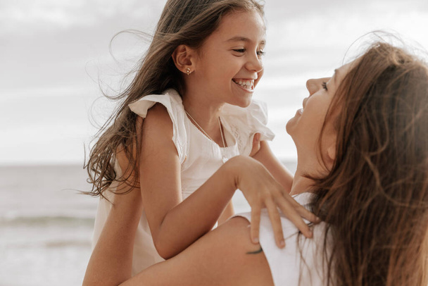 Side view of happy young woman with long dark hair in white dress, embracing adorable positive little daughter and looking at each other on seashore against cloudy sky - Фото, изображение