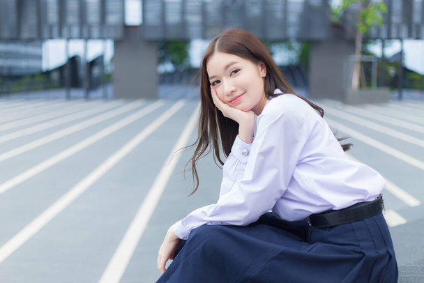 Όμορφη Ασιάτισσα μαθήτρια λυκείου με σχολική στολή με σιδεράκια στα δόντια κάθεται και χαμογελάει με αυτοπεποίθηση ενώ κοιτάζει την κάμερα χαρούμενη με το κτίριο στο βάθος. - Φωτογραφία, εικόνα