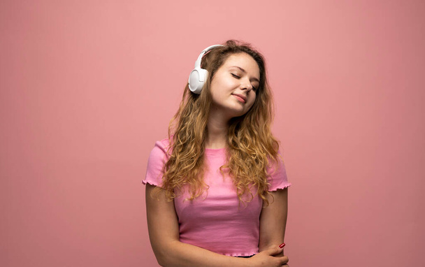 Portrait of charming curly woman in white massive headphones listening to music on isolated pink background. Curly girl in t-shirt smiles - Photo, Image