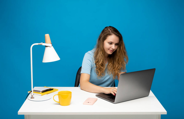 Happy young freelancer curly woman in a blue t-shirt working with a laptop computer and speaking with a client or friends with messengers. Working on a project. Freelance worker - Photo, image