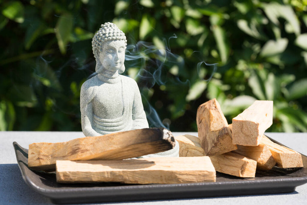 Incense burning. Burning palo santo sticks with Buddha statue against green leaves as background under the sunlight. Zen concept, meditation and spiritual practices. Selective focus, focus on statue - Foto, Imagen