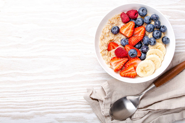 Oatmeal porridge with fruit and berries in bowl with spoon on white wooden background table top view, homemade healthy breakfast cereal with strawberry, banana, blueberry, raspberry. Space for text - Foto, imagen