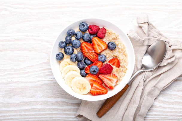 Oatmeal porridge with fruit and berries in bowl with spoon on white wooden background table top view, homemade healthy breakfast cereal with strawberry, banana, blueberry, raspberry. Space for text - Photo, Image