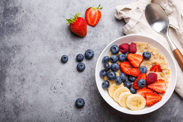 Oatmeal porridge with fruit and berries in bowl with spoon on gray stone background table top view, homemade healthy breakfast cereal with strawberry, banana, blueberry, raspberry. Space for text - Photo, Image