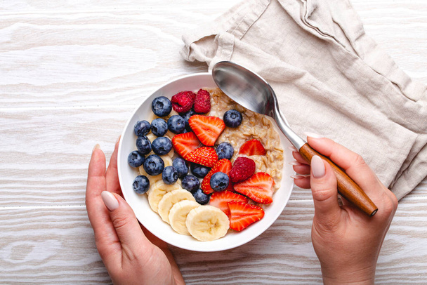 Top view of female hands holding bowl with oatmeal porridge with fruit and berries and spoon on white wooden background table top view, woman eating homemade healthy breakfast cereal - Photo, Image
