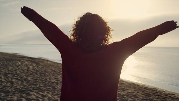 Takaisin tarkastella rento nainen nostaa kätensä rannalla. Romanttinen tyttö tuntee vapautta merellä. Naishipsteri kävelee hiekkarannalla. Iloinen nainen nauttimassa merimaisemasta auringonnousun valossa. Voittajan käsite - Valokuva, kuva