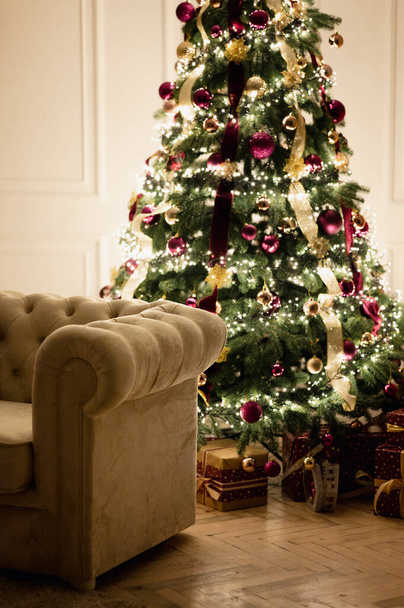 Christmas Tree is Decorated with Purple and Gold Toys at Night. Chesterfield Sofa in Foreground. White Parquet. - Foto, immagini