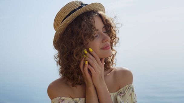 Seductive woman closed eyes enjoy sunlight on beautiful seacoast close up. Portrait of attractive curly girl wearing straw hat gentle touching face. Relaxed young lady posing on sunny summer beach. - Photo, Image