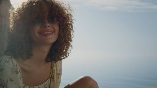 Söpö positiivinen tyttö nauraa näköinen kamera lähellä rauhallinen sininen meri lähikuva. Muotokuva onnellinen nuori nainen istuu hiekkarannalla aurinkoinen kesäpäivä. Curly houkutteleva malli aurinkolasit laskettu pää hymyillen.  - Valokuva, kuva