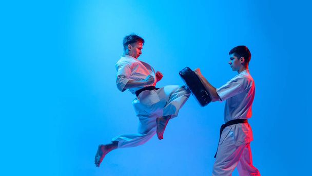 Studio shot of sports training of two karatedo fighters in doboks isolated on blue background in neon. Concept of combat sport, challenges, skills. Sportsmen practicing base technique - Foto, imagen