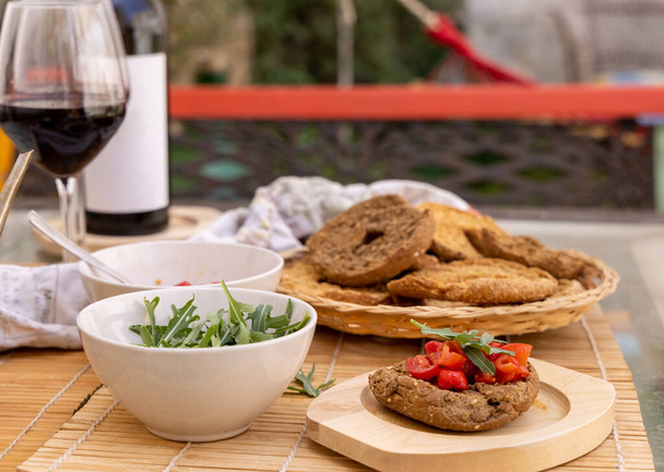 Italian appetizer Friselle with cherry tomatoes, oil and rucola served outdoor in with glasses of red wine. Tipical Apulian Dried bread, made in Salento. Local cousine. Vegetarian food - Φωτογραφία, εικόνα