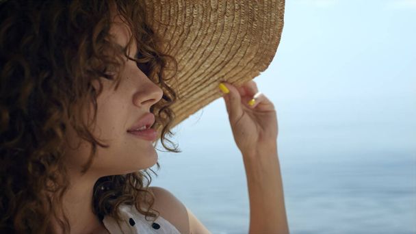 Portrait de luxueuse femme bouclée portant un chapeau large en paille sur une plage ensoleillée. Attrayant confiant fille regardant caméra séduisante détente sur le rivage gros plan. Sexy jeune modèle profiter de la lumière du soleil d'été. - Photo, image