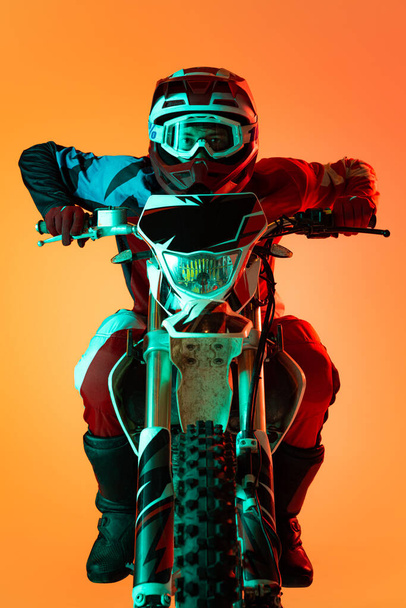 Portrait of young concentrated man, biker developing speed on motorbike isolated over orange studio background in neon light. Concept of motosport, speed, action, hobby, competition. Copy space for ad - 写真・画像