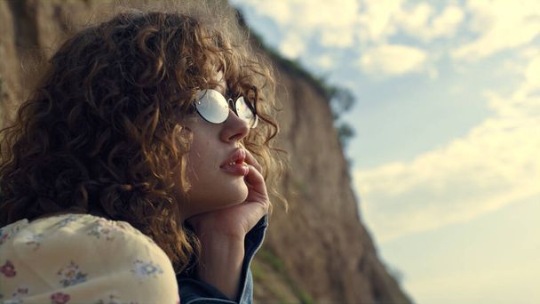 Dreamy fashionable woman portrait sitting alone wearing sunglasses on sand seashore. Attractive curly girl turning face to camera close up. Tranquil young lady enjoy last evening rays on empty beach. - Photo, Image