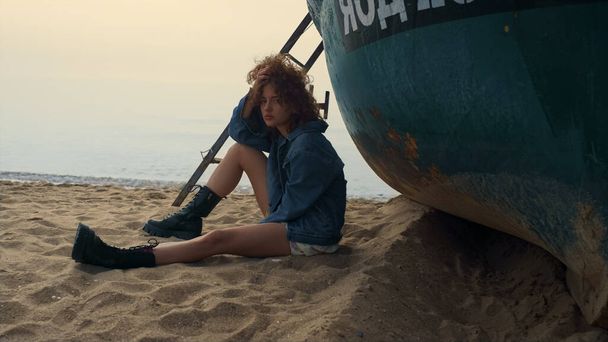 Smutná mladá žena sedí na pláži poblíž staré lodi zamračené ráno. Atraktivní stylová dívka pózující na pobřeží dotýkající se krásných kudrnatých vlasů. Rozrušená krásná dáma, co vypadá jako kamera. Volný čas v přírodě podzimní den. - Fotografie, Obrázek