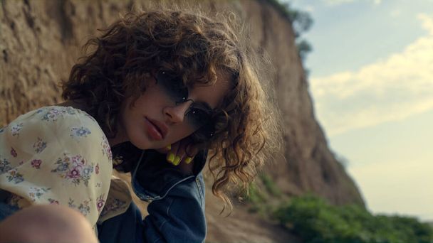 Portrait of seductive girl wearing stylish sunglasses in front sand hill. Attractive curly lady posing sitting beautiful seashore in fashion clothes. Young gorgeous woman looking camera confidently. - Foto, Bild