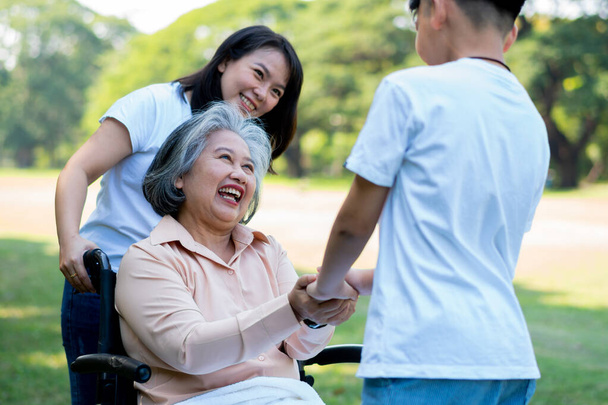 Ευτυχισμένη ηλικιωμένη Ασιάτισσα γιαγιά χρησιμοποιεί αναπηρικό καροτσάκι με την κόρη της και το εγγόνι της στο πάρκο, ο εγγονός ήρθε να επισκεφθεί ηλικιωμένη γιαγιά και να κρατήσει το χέρι. Έννοια της ευτυχισμένης οικογένειας, καλή σχέση μαζί - Φωτογραφία, εικόνα