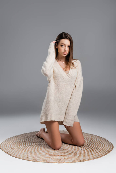 young woman in sweater and with bare legs kneeling on carpet on grey background - 写真・画像