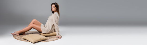 повна довжина жінки з тонкими голими ногами, сидячи на килимі в м'якому светрі на сірому фоні, банер
 - Фото, зображення