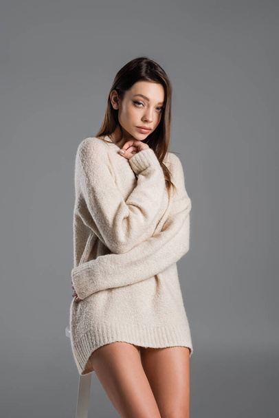 pretty brunette woman in warm sweater hugging herself and looking at camera isolated on grey - Foto, imagen