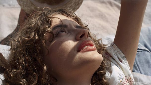 Beautiful relaxed woman hiding face from sunlight by hand close up. Portrait of attractive curly girl lying on sand beach summer holiday. Cute carefree lady enjoy tranquil rest on sunny seashore. - Fotó, kép