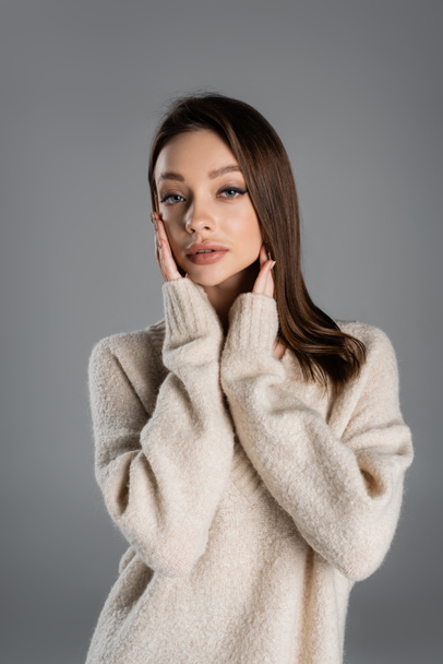 pretty woman in cozy sweater touching face while looking at camera isolated on grey - Foto, Imagen