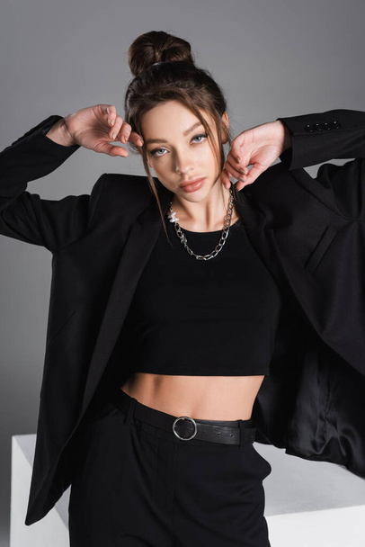 sensual woman in black crop top and blazer posing with hands near face isolated on grey - Фото, изображение